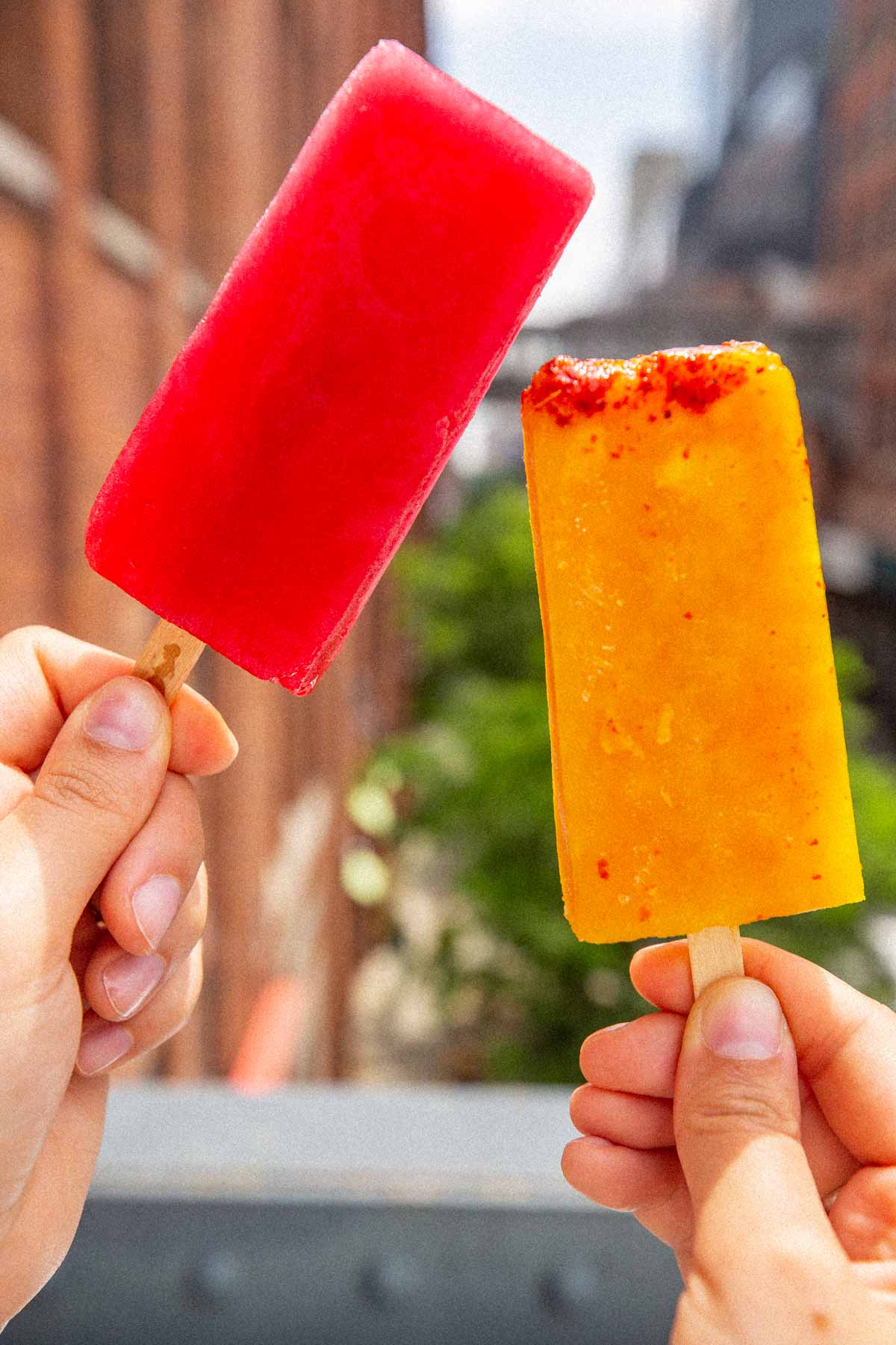 Two hands holding up a bright red and an orange chili-dusted paleta with the High Line and a brick building behind them
