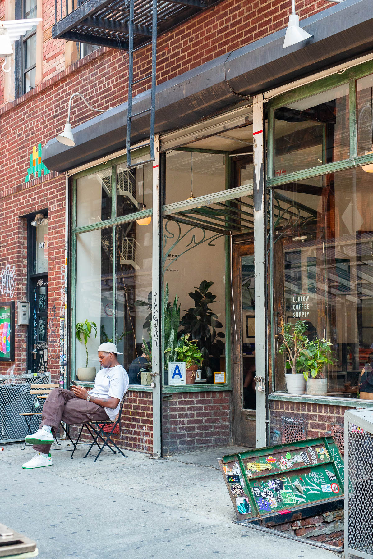 Man sitting at a bistro table outside of Ludlow Coffee Supply, whose windows are filled with plants