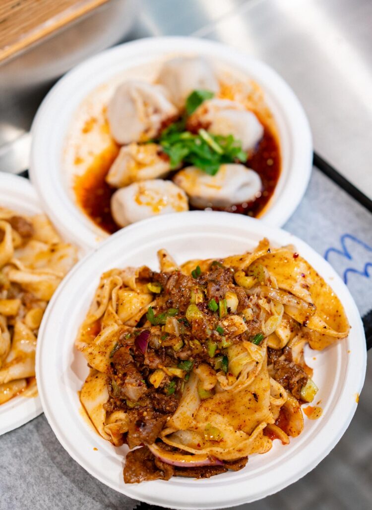 A foam plate of Xi'an Famous Foods spicy cumin lamb hand-ripped noodles beside a plate of pork wontons in red chili oil at a Flushing food stall