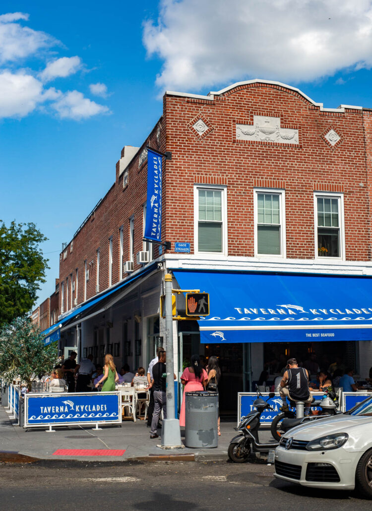 The blue-awninged corner of Taverna Kyclades on Ditmars Boulevard in Astoria, with diners at sidewalk tables and a small crowd waiting outside